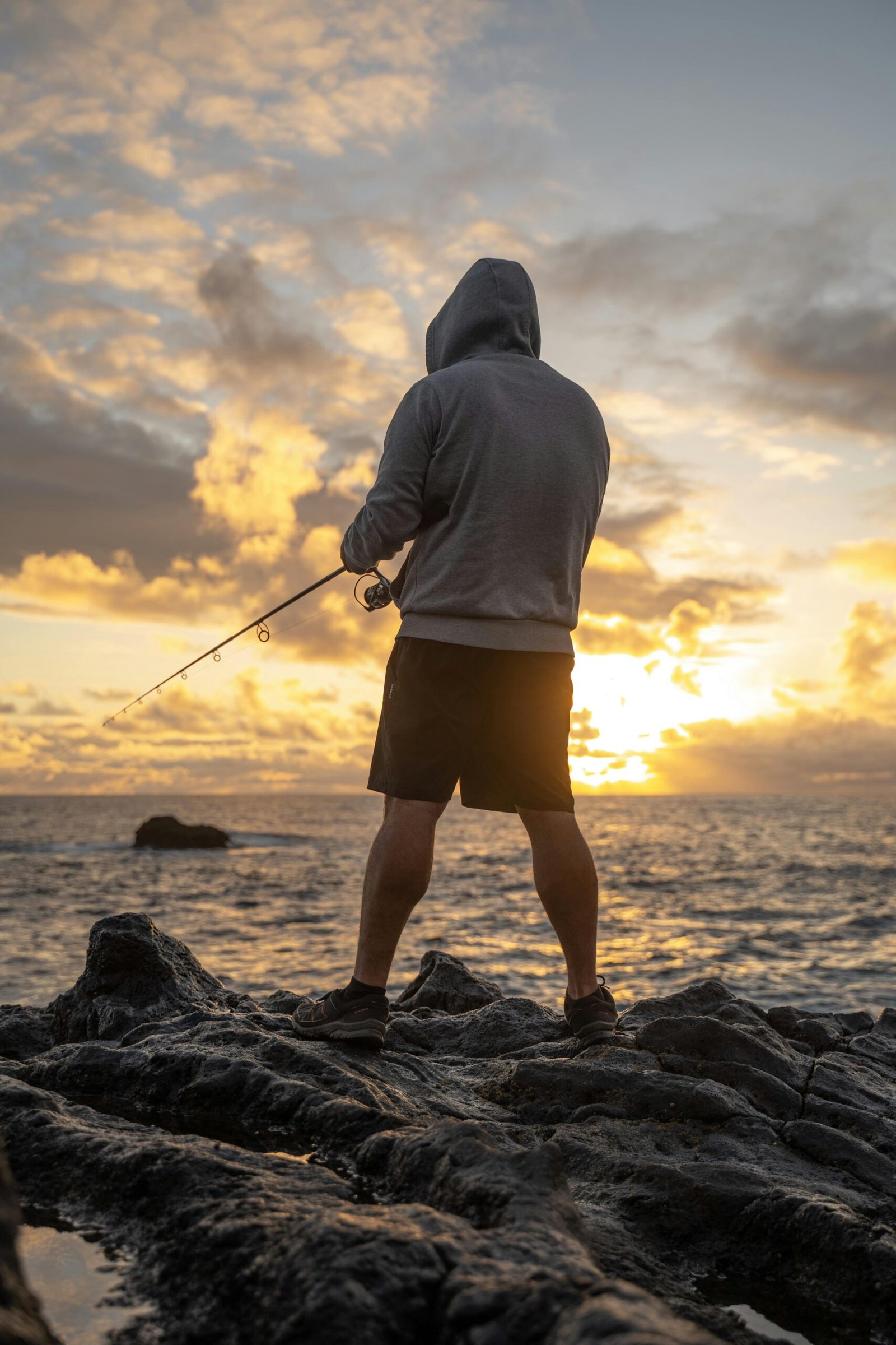 Back view of a man fishing on a rocky coast at sunset, enjoying outdoor recreation.