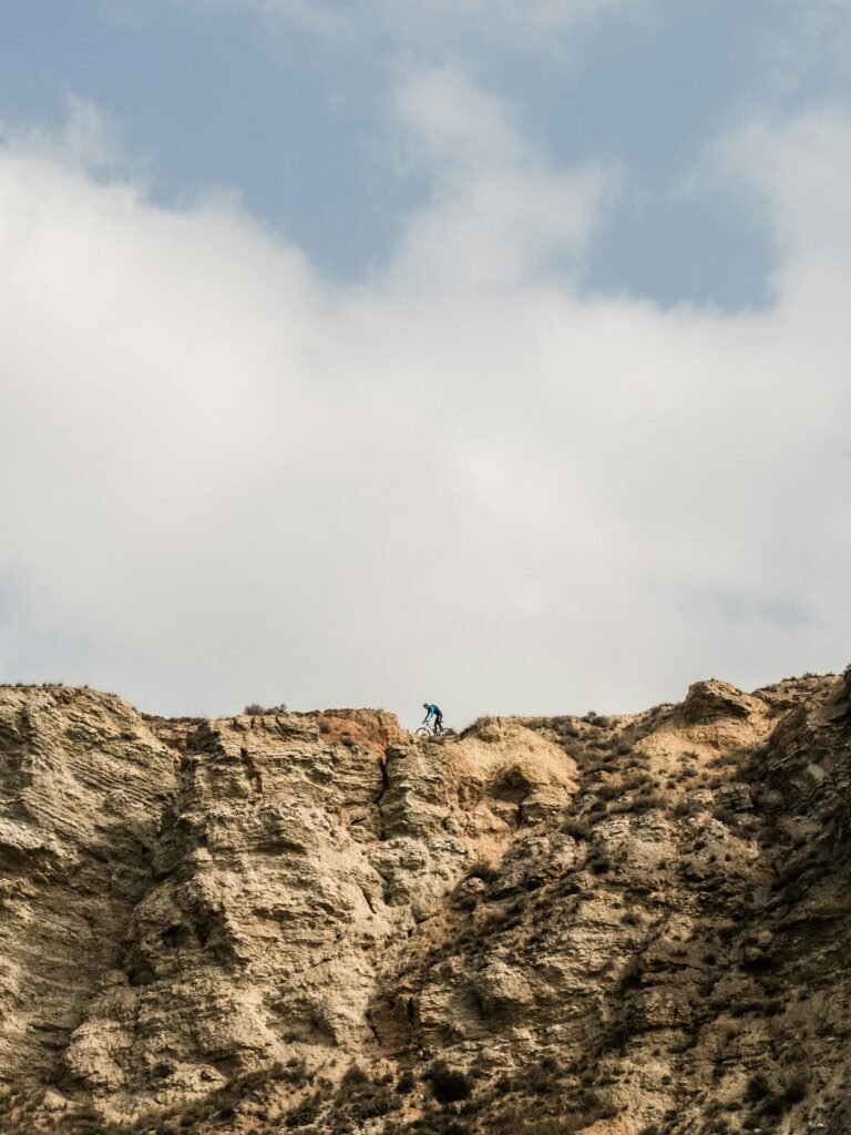 A lone mountain biker navigates the rocky cliffs with white clouds above.
